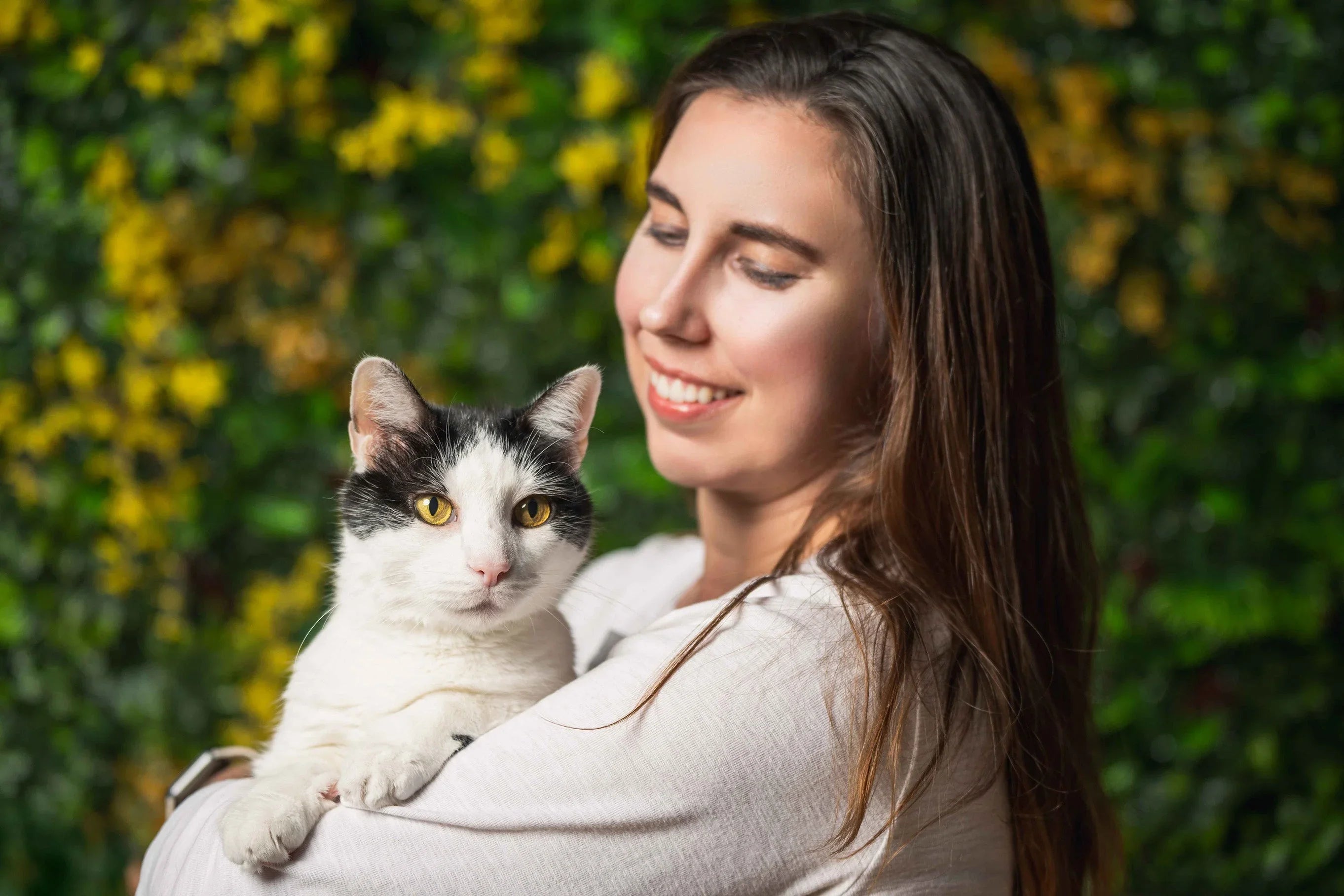 Smiling woman holding black and white cat with yellow eyes against green leafy background